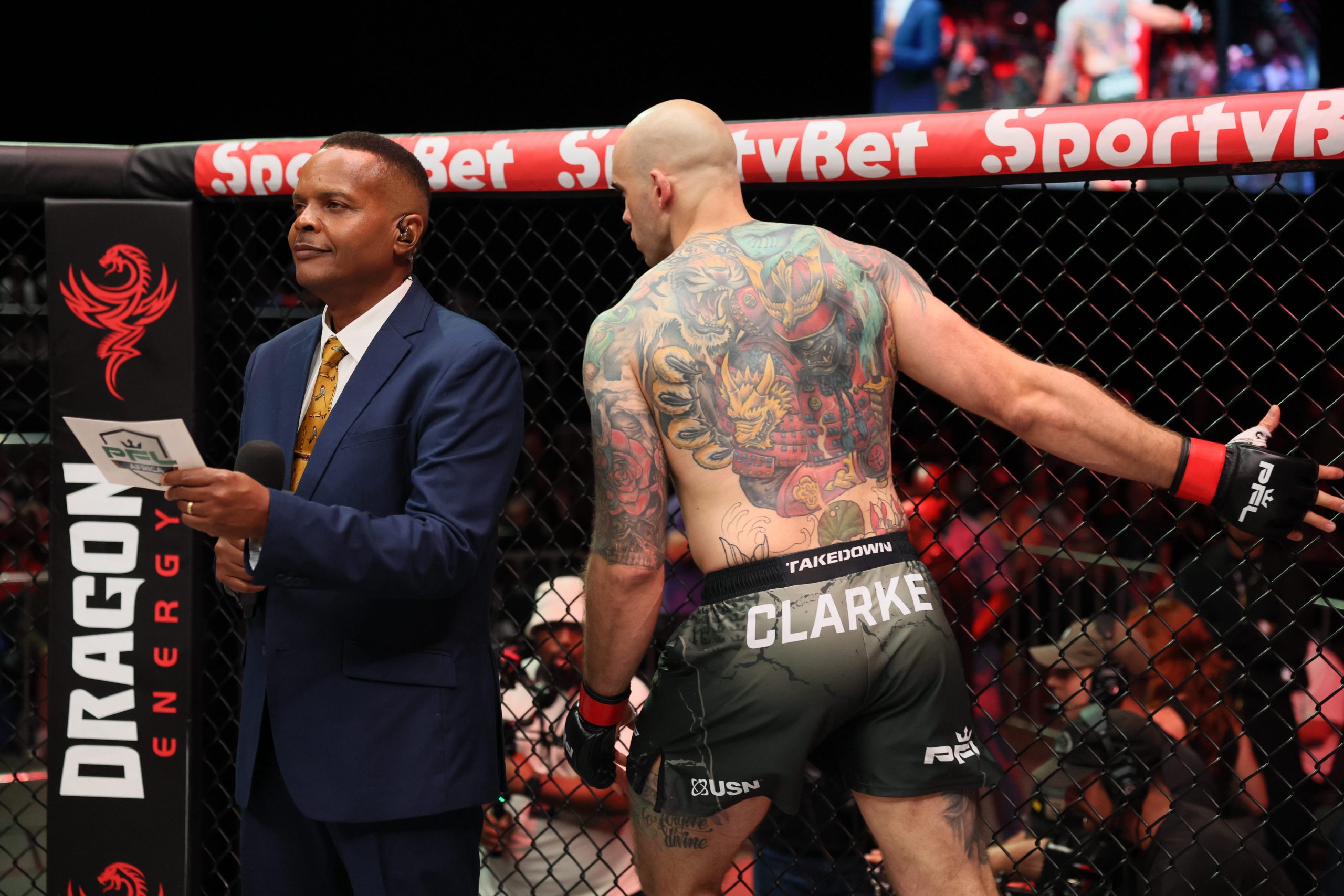 Announcer in a suit with a PFL Africa cue card beside fighter Clarke at the cage fence